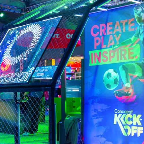 Fotografía del estand promocional de Concacaf KICK-OFF en un evento de videojuegos. En el lado izquierdo se aprecia una estructura metálica con red iluminada por luces neón en tonos azul y verde, coronada por el logotipo oficial de Concacaf. Del lado derecho se observa una lona publicitaria vertical con fondo azul y el mensaje "CREATE. PLAY. INSPIRE." sobre la imagen de una jugadora de fútbol pateando un balón. En la esquina inferior derecha resalta el logotipo de Concacaf KICK-OFF.