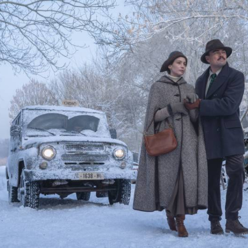 Escena invernal con dos personajes vestidos con ropa de época, de pie frente a un vehículo antiguo en un camino nevado rodeado de árboles.