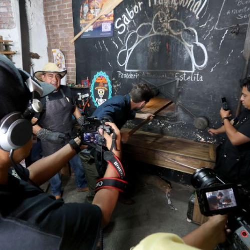 Fotografía de detrás de cámaras durante el rodaje del documental 'Entre 2 Mundos' en una panadería artesanal. En el centro, el actor y conductor Arap Bethke viste una camisa azul y manipula una pala de madera para introducir pan en un gran horno de ladrillo tradicional marcado con la leyenda 'Sabor Tradicional Panadería Estelita'. Al lado del horno, un panadero con delantal y sombrero de paja observa el proceso. En primer plano y a los costados se observan camarógrafos y fotógrafos sosteniendo cámaras profesionales registrando la escena.