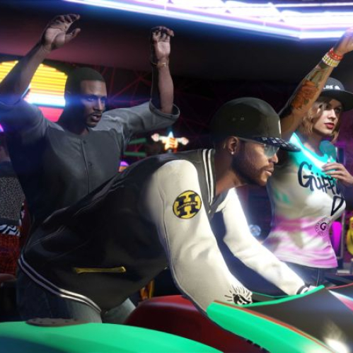 Captura de pantalla de un grupo de personajes dentro de un establecimiento de maquinitas recreativas en GTA Online. En primer plano, un hombre joven de espaldas con gorra negra y sudadera universitaria blanca y negra con un logotipo amarillo en la manga opera un simulador de carreras de motocicletas de color verde. Detrás de él, un hombre afroamericano con las manos levantadas en señal de festejo y una mujer con tatuajes en el brazo y gorra negra observan la partida con entusiasmo en un entorno iluminado por luces de neón moradas y pantallas de juegos arcade como Badlands Revenge II.