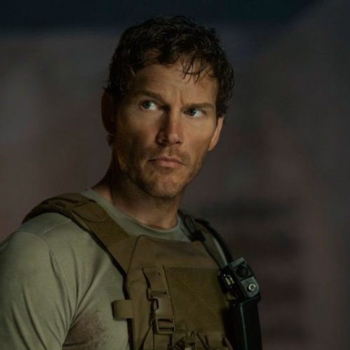 Fotografía promocional en plano medio corto del actor Chris Pratt caracterizado como el comandante James Reece. Viste un chaleco táctico color caqui sobre una playera beige con manchas de tierra. Tiene el cabello ligeramente despeinado, rostro serio con barba corta y mira de perfil hacia su derecha en un entorno con iluminación cálida y tenue.