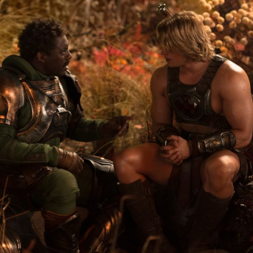 Fotografía fija de la película "Amos del Universo". En un entorno natural de vegetación alta y tonos otoñales, dos personajes conversan de perfil. A la izquierda, Duncan/Man-At-Arms, interpretado por Idris Elba con tez morena, viste una armadura metálica de tonos oscuros y bronce con acentos verdes en las mangas. A la derecha, el príncipe Adam/He-Man, interpretado por Nicholas Galitzine con cabello rubio semilargo, viste una armadura pectoral metálica con detalles ornamentales y sostiene sus manos entrelazadas mientras observa fijamente a su compañero.