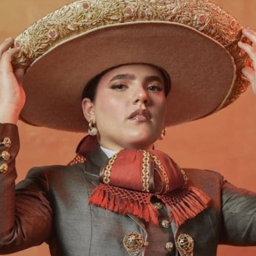 Fotografía de estudio de la cantante Camila Fernández posando con un elegante traje de chargris o gris oscuro con detalles y botonadura dorada de estilo tradicional. La artista porta un sombrero de charro de ala ancha en tono beige con bordados dorados, el cual sostiene con ambas manos a la altura de la cabeza, mientras luce una corbata de moño en tonos rojos y una expresión segura mirando directamente a la cámara sobre un fondo cálido difuminado.