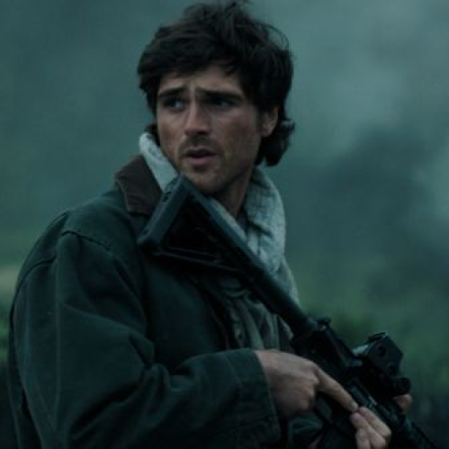 Fotografía en formato horizontal de una escena de la película ambientada en un entorno exterior y brumoso. En el centro, el actor Jacob Elordi aparece caracterizado como Hig, luciendo cabello castaño despeinado, barba corta y una bufanda gris con chamarra verde oliva. Sostiene un rifle de asalto de perfil mientras mira con preocupación hacia la izquierda, con una pequeña hoguera encendida al fondo.