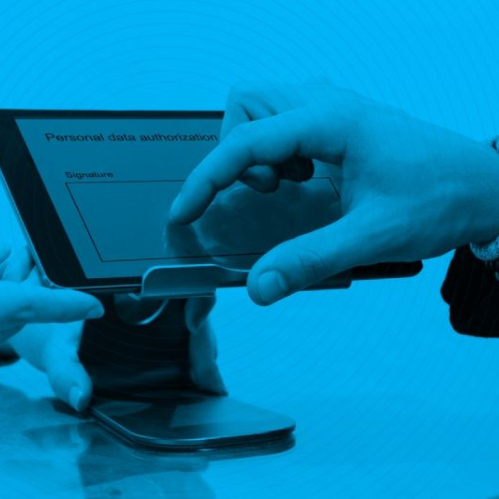 Fotografía en tono monocromático azul que muestra a una persona utilizando su dedo índice para firmar digitalmente un documento de autorización de datos personales en la pantalla de una tablet montada en un soporte sobre un escritorio.