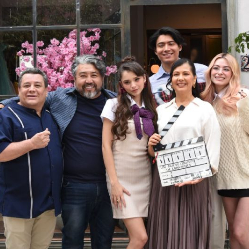 Elenco y equipo de producción sosteniendo la claqueta oficial durante el pizarrazo de inicio de grabaciones de 'Un Nuevo Contrato de Corazones'. En el centro destaca la actriz Farah Justiniani vistiendo un uniforme beige con moño morado junto a la actriz Aida Morales sosteniendo la claqueta blanca con los datos del rodaje. Al fondo sonríe el actor Tristan Maze. A los lados se observan actores del elenco como Roberto Tello y miembros de producción en una locación urbana con decoración de cerezos rosados.