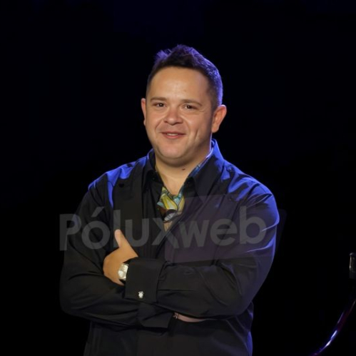 Fotografía de Rodrigo de la Cadena posando sonriente con los brazos cruzados junto a un piano de cola en un escenario con iluminación azul y fondo oscuro durante la presentación del evento.
