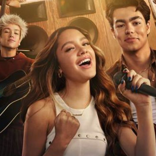 Fotografía promocional en plano medio del elenco juvenil de la película Camp Rock 3. En el centro de la imagen se encuentra una joven cantante con cabello castaño largo y ondulado sosteniendo un micrófono negro profesional mientras sonríe. Viste una blusa blanca sin mangas con detalles metálicos. A la izquierda, en segundo plano, un joven de cabello rubio platinado despeinado toca una guitarra acústica luciendo una sudadera guinda. A la derecha, otro joven sonriente con cabello castaño peinado hacia atrás sostiene una guitarra eléctrica luciendo una camisa de franela a cuadros en tonos cafés. Al fondo se aprecian bocinas y equipo de sonido sobre una pared de madera.