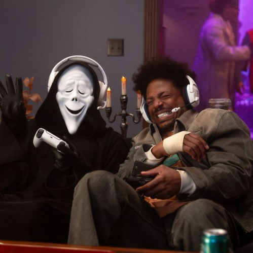 Un hombre sentado en un sillón junto a un personaje con la túnica negra y máscara sonriente de Ghostface. Ambos sostienen mandos de consola de videojuegos y llevan auriculares blancos con micrófono integrados, riendo en una habitación decorada con velas sobre candelabros.