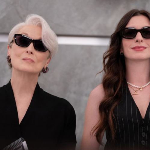 Dos mujeres con atuendos negros y gafas oscuras posan en un entorno elegante de mármol gris.