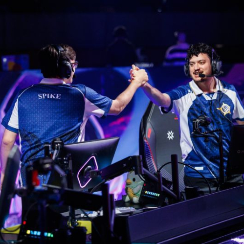 Dos jugadores de esports celebran en un escenario competitivo, vestidos con uniformes de equipo y rodeados de iluminación azul y púrpura.