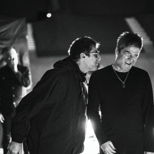 Fotografía en blanco y negro en plano medio durante el backstage de un concierto. Se observa a Liam Gallagher en el centro, vistiendo chamarra de cuello alto y anteojos oscuros, riendo mientras se inclina hacia su hermano Noel Gallagher, quien viste una playera de manga larga, cadena al cuello y sonríe ampliamente. Al fondo a la izquierda se distingue otra figura en la penumbra.