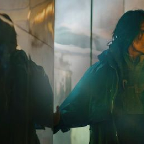 Fotografía cinematográfica en plano medio con tonalidades frías y azuladas que muestra a la actriz Michelle Yeoh interpretando a Olwen. Luce una chaqueta larga de invierno color azul verde con abrigo grueso y el cabello castaño oscuro despeinado a la altura de los hombros. Apoya su mano sobre una pared o cristal brillante con reflejos metálicos mientras mira con expresión seria hacia el lado derecho de la toma.
