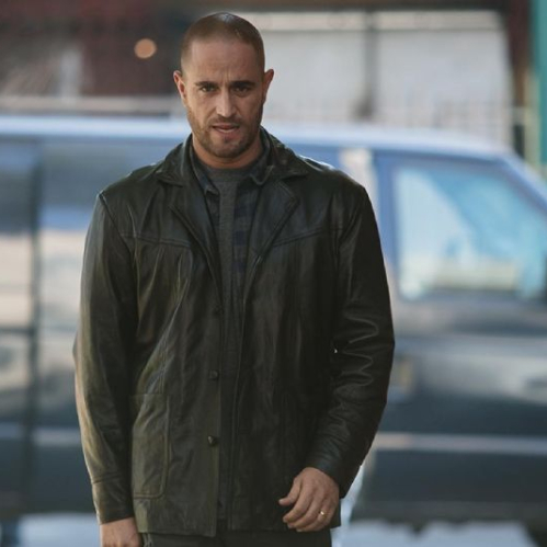 Plano medio del actor Michel Brown interpretando al detective Alejandro Falco. Viste una chaqueta de piel negra abierta sobre una playera azul marino con rayas horizontales oscuras. Luce el cabello rapado, barba corta y una expresión seria y concentrada mientras camina al aire libre en una calle urbana con una camioneta oscura estacionada de fondo.