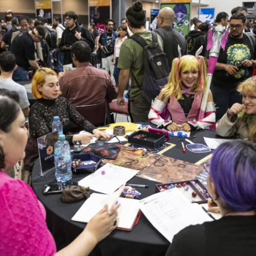 Jugadores reunidos alrededor de una mesa en un evento concurrido, con materiales de rol y un ambiente lleno de actividades.