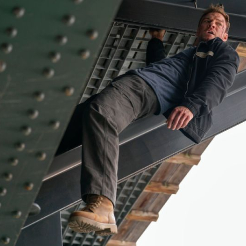 Fotografía de acción en plano medio con ángulo contrapicado. El actor Alan Ritchson interpretando a Jack Reacher cuelga de la estructura metálica inferior de un puente o viga industrial de acero con remaches. Viste una chamarra oscura con playera azul marino, pantalones cargo oscuros y botas de trabajo marrones; se aprecian raspones y heridas de combate en su cara y frente mientras mira hacia un lado con tono de urgencia.