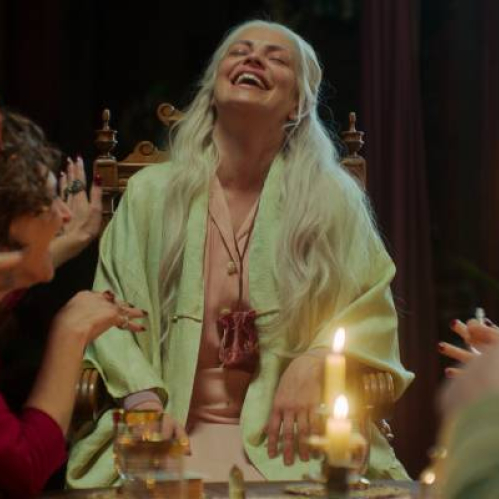 Un grupo de personas comparte una velada íntima alrededor de una mesa iluminada por velas, mientras una mujer mayor ríe con energía en el centro de la escena.