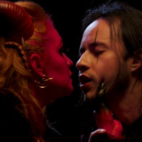 Dos intérpretes en una escena dramática con iluminación oscura y tonos rojizos, uno de ellos con cuernos estilizados, creando una atmósfera intensa y teatral.