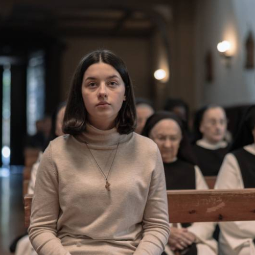 Persona sentada en un espacio religioso, acompañada por figuras vestidas con hábitos, evocando introspección y contemplación.