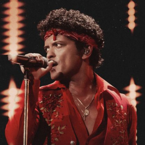 Fotografía en primer plano de Bruno Mars cantando frente a un micrófono sobre el escenario con camisa roja desabotonada, paliacate rojo en la cabeza y luces de fondo.