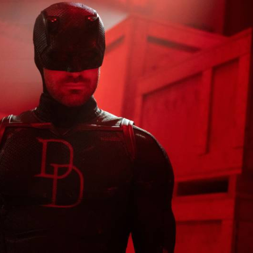 Figura en traje oscuro con máscara y el emblema “DD” en el pecho, iluminado por tonos rojos en un entorno industrial.