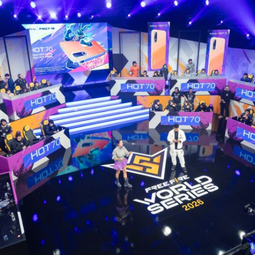 Fotografía del escenario principal de un torneo de esports de la Free Fire World Series Brazil 2026. La tarima cuenta con iluminación morada y azul dramática, una pantalla gigante en el centro que muestra el logotipo de Free Fire World Series 2026 y promocionales del smartphone Infinix HOT 70. En los laterales hay estaciones de juego elevadas con computadoras y monitores donde compiten los jugadores, mientras que en el centro del escenario dos presentadores conducen el evento.