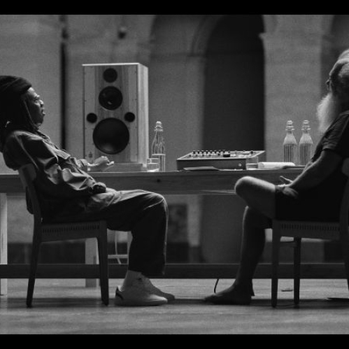 Fotografía en blanco y negro donde se aprecia a Jay-Z y al productor Rick Rubin sentados frente a frente en sillas de madera a los extremos de una mesa larga. Al centro de la mesa se observa un altavoz de estudio y equipo de sonido. Jay-Z, del lado izquierdo, luce vestimenta casual con gorro y calzado blanco, mientras Rick Rubin, del lado derecho, viste playera oscura, shorts y luce su característica barba larga y canosa.