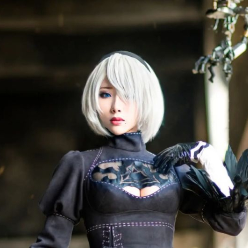 Fotografía de la cosplayer internacional Hane Ame caracterizada como el personaje 2B del videojuego NieR: Automata. Luce peluca blanca corta, vestido negro bordado con detalles en el pecho y plumas en las mangas, mientras sostiene una espada en un entorno industrial.
