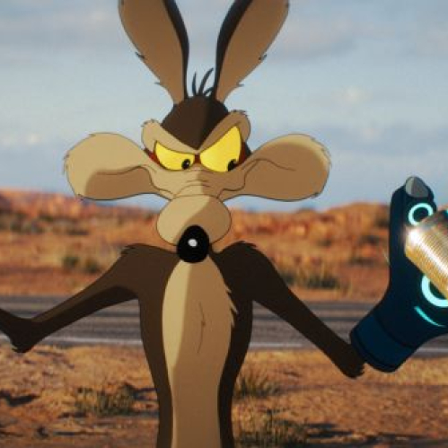 Fotografía promocional en plano medio al aire libre con fondo de paisaje desértico de cañones al atardecer. En el centro, el personaje animado del Coyote se muestra parado sobre la carretera con una expresión seria y concentrada mirando de reojo hacia la derecha. Porta un guante tecnológico de color azul con círculos luminosos en su mano izquierda, sosteniendo en la palma una pequeña lata metálica cilíndrica.
