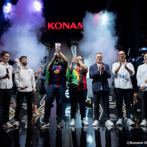 Escenario principal del eFootball World Festival en Bangkok lleno de luces dramáticas y humo blanco. En el centro del escenario, dos jugadores de esports vistiendo camisetas deportivas de fútbol y banderas de Brasil sobre los hombros levantan trofeos de cristal transparente sobre sus cabezas. A sus lados, organizadores con micrófonos, ejecutivos y la exfigura del fútbol Wayne Rooney vestido con saco oscuro y playera blanca aplauden el momento de la premiación. Al fondo se observa la pantalla gigante con el logotipo rojo de KONAMI.