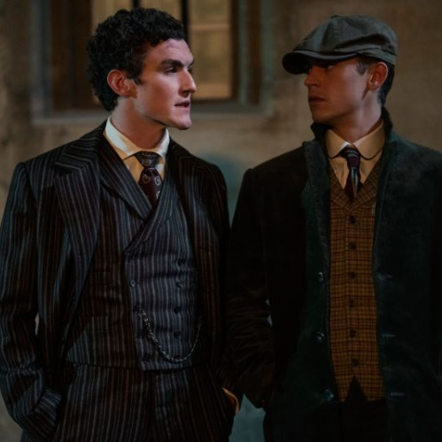 Fotografía promocional de época en plano medio de Hero Fiennes Tiffin y Dónal Finn parados al aire libre frente a una construcción clásica iluminada de noche. A la izquierda, Dónal Finn viste un traje de tres piezas a rayas oscuras con corbata estampada y cadena de reloj de bolsillo. A la derecha, Hero Fiennes Tiffin lleva un abrigo verde oscuro sobre un chaleco a cuadros, camisa blanca, corbata y una boina plana de época, mientras observa fijamente a su compañero. Crédito de foto: Daniel Smith.