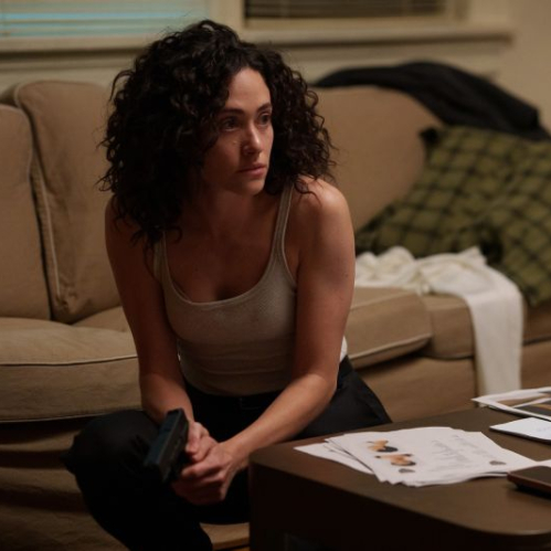 La actriz Emmy Rossum caracterizada como la agente del FBI Alice Black en la serie 'Furia'. Se encuentra agachada vestida con una playera de tirantes clara y pantalón oscuro, sosteniendo una pistola con la mano derecha frente a una mesa de centro que contiene expedientes, fotografías y teléfonos móviles. Al fondo se observa un sillón color beige con ropa y un cojín.