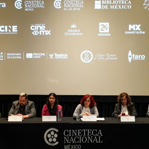 Fotografía de una conferencia de prensa en una sala de cine de la Cineteca Nacional México. Frente a una pantalla donde se proyectan los logotipos de la Secretaría de Cultura, Cineteca Nacional, IMCINE y Canal 22, seis ponentes se encuentran sentados detrás de una mesa de presídium cubierta con mantelería negra con el logotipo del recinto en blanco. Los ponentes cuentan con micrófonos y hojas sobre la mesa.