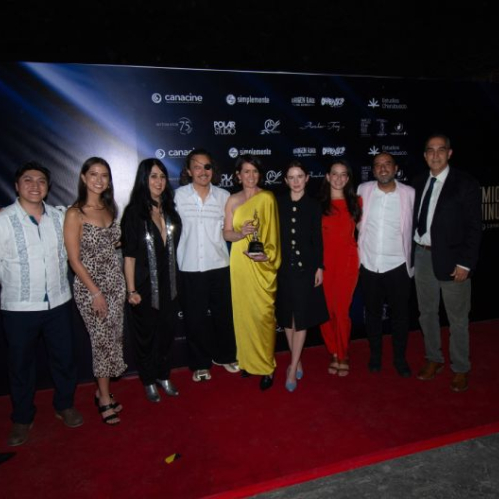 Fotografía grupal sobre alfombra roja durante la entrega de los Premios Luminus CANACINE. Al centro destaca una mujer con vestido largo color amarillo sosteniendo la estatuilla dorada, rodeada por integrantes de los equipos de producción, talento y directivos, vestidos con traje formal, vestidos de noche y camisas de gala frente a un muro de prensa con logotipos del evento.