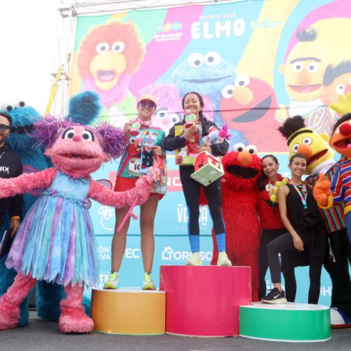 Fotografía en el podio de premiación donde dos corredoras posan con sus trofeos acompañadas por las botargas de Abby Cadabby, Elmo, Beto, Enrique y los presentadores del evento.