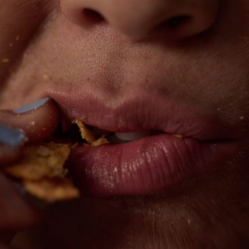 Plano detalle en acercamiento de la parte inferior del rostro de una persona comiendo. Se observan de cerca los labios entreabiertos, la barbilla y la parte inferior de la nariz mientras la mano con uñas pintadas de color azul claro introduce trozos de galleta o alimento crujiente a la boca, dejando migajas sobre los labios y la piel.