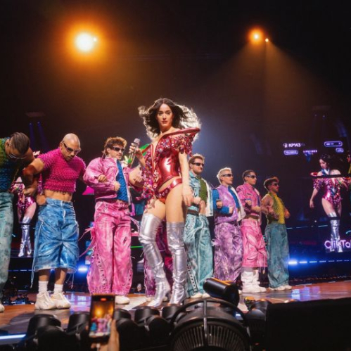Fotografía en plano general de Katy Perry actuando en vivo en un escenario iluminado por luces cálidas. La cantante viste un body metálico rojo con botas altas plateadas mientras sostiene un micrófono y camina al frente. A sus espaldas la acompañan ocho bailarines con atuendos coloridos en tonos rosa, azul y verde posando en línea. Al fondo se observa la pantalla del escenario y en el primer plano inferior las luces de los dispositivos del público.