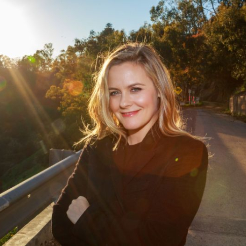 Retrato al aire libre en plano medio de Alicia Silverstone sonriendo a la cámara al atardecer. La actriz luce cabello rubio peinado con ondas suaves sobre los hombros y viste un saco o abrigo negro mientras posa con los brazos cruzados. Al fondo se observa una carretera de montaña iluminada por un destello cálido de luz solar.