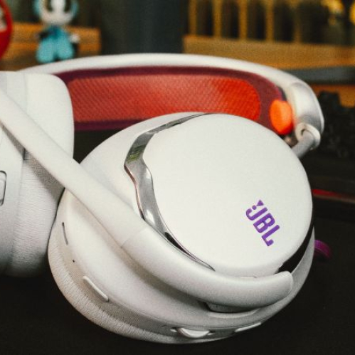 Audífonos para videojuegos JBL Quantum 950 de color blanco con detalles morados y naranja sobre un escritorio junto a un teclado gamer retroiluminado.