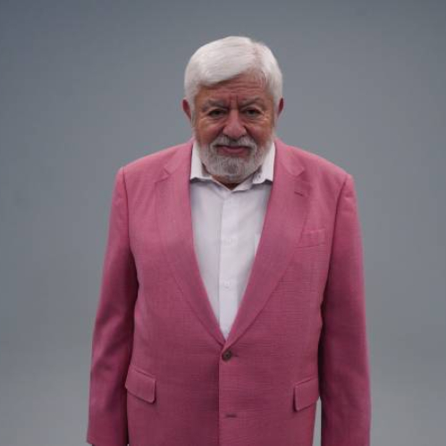 Persona de cabello blanco y barba posando con un saco rosa brillante frente a un fondo gris neutro, transmitiendo presencia y personalidad.