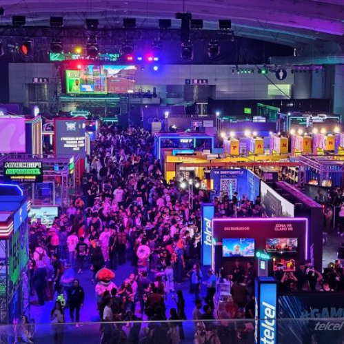 Vista panorámica desde una planta elevada del piso de exhibición de Telcel GAMERGY 2026 en Expo Santa Fe, repleto de asistentes caminando entre pasillos iluminados con luces moradas, azules y de neón, rodeados por pabellones de marcas como Telcel Infinitum, Minecraft Experiencia, Just Dance y zonas de VR.