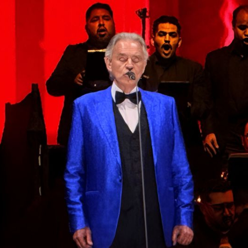 Un intérprete de traje azul canta frente a un coro vestido de negro bajo iluminación roja intensa.