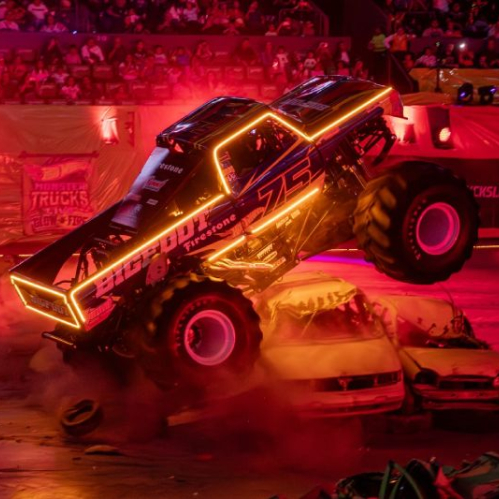 Un monster truck iluminado con luces de neón aplasta dos autos en una arena llena de público.