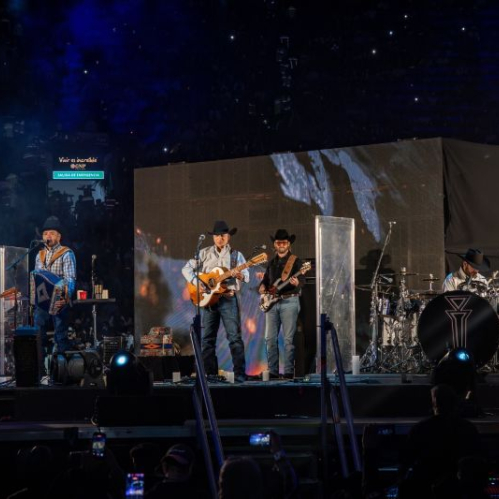 Banda norteña tocando en un escenario con pantallas y luces en un recinto masivo.