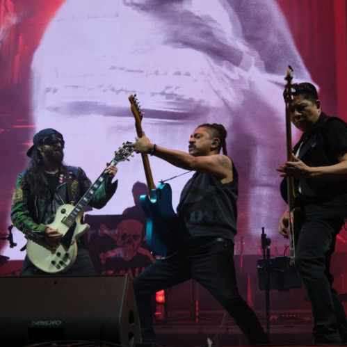 Tres músicos de Panteón Rococó tocan en un escenario iluminado con luces rojas y una gran pantalla al fondo, en la que se proyecta una imagen en blanco y negro, capturando la intensidad de un show en vivo.