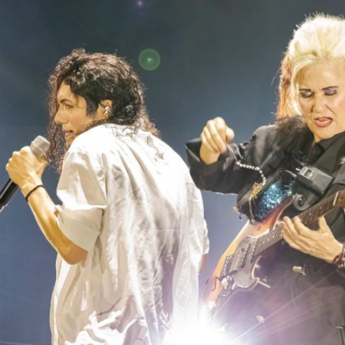 El intérprete del tributo cantando en un micrófono blanco junto a la guitarrista Jennifer Batten tocando su guitarra eléctrica iluminados por luces de escenario.