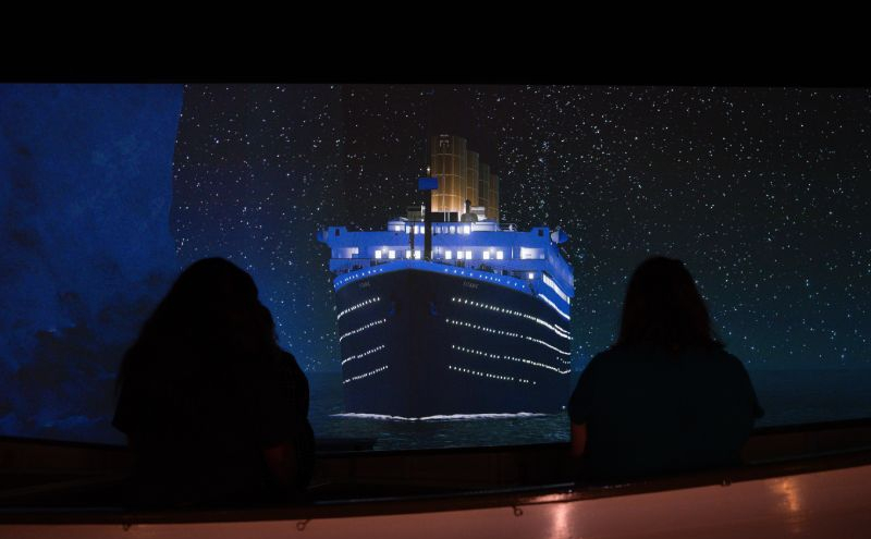 Espectadores sentados observando una gran proyección frontal que muestra la proa del Titanic navegando bajo un cielo estrellado.
