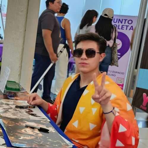 Persona con gafas oscuras y atuendo inspirado en un kimono amarillo sentado en una mesa llena de ilustraciones estilo anime durante un evento concurrido.