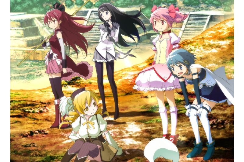 Arte oficial de las protagonistas del ciclo "Camino hacia Walpurgisnacht Rising" de Madoka Magica. Ilustración oficial en formato horizontal al aire libre con ruinas de piedra y vegetación de fondo bajo la luz del sol. Aparecen las cinco chicas mágicas principales del anime con sus trajes de combate. A la izquierda, Kyoko Sakura vestida de rojo sostiene su lanza de madera; detrás, Homura Akemi con uniforme escolar blanco y medias oscuras; al centro, Mami Tomoe sentada en el suelo vestida de amarillo; a la derecha, Madoka Kaname en su vestido rosa con moños; y en primer plano a la derecha, Sayaka Miki inclinada sonriendo con su capa blanca y traje azul. Abajo al centro asoma la cabeza blanca de Kyubey.
