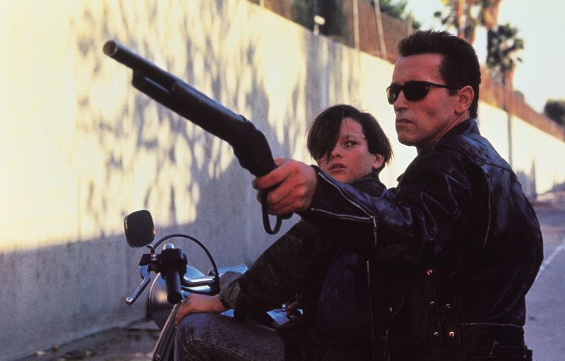 Arnold Schwarzenegger y Edward Furlong en "Terminator 2: El juicio final". Fotografía en formato horizontal de una escena en exterior durante el día. A la derecha, el actor Arnold Schwarzenegger caracterizado como el T-800 viste una chamarra de cuero negra y gafas de sol oscuras mientras sostiene una escopeta recortada hacia el frente con la mano derecha. Detrás de él, sobre la motocicleta, se observa al actor Edward Furlong en el papel de John Connor, mirando de perfil hacia la escopeta. Al fondo se distingue una barda de concreto clara y palmeras bajo la luz del sol.